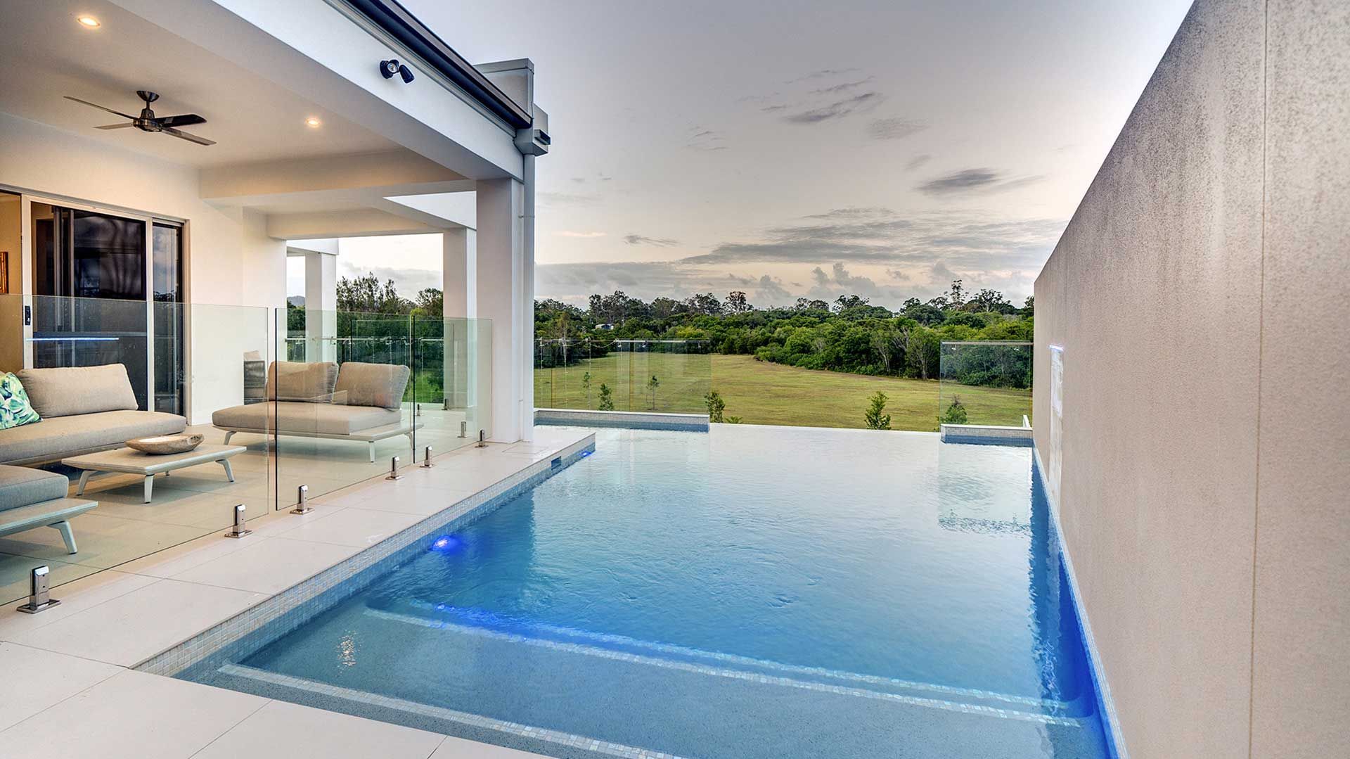 An infinity pool with a blue hue, patio, and a view of fields under a cloudy sky.