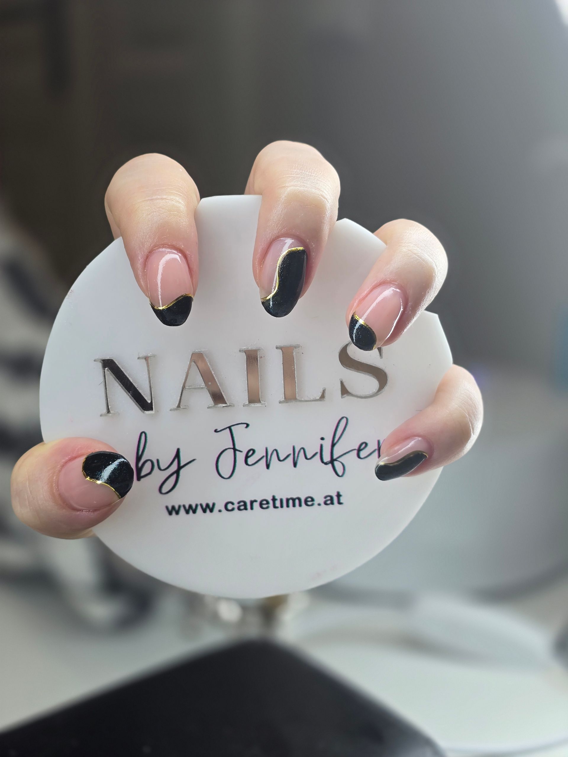 Eine Hand hält eine weiße, runde Karte mit der Aufschrift „NAILS by Jennifer“ und ihre Fingernägel sind schwarz lackiert.