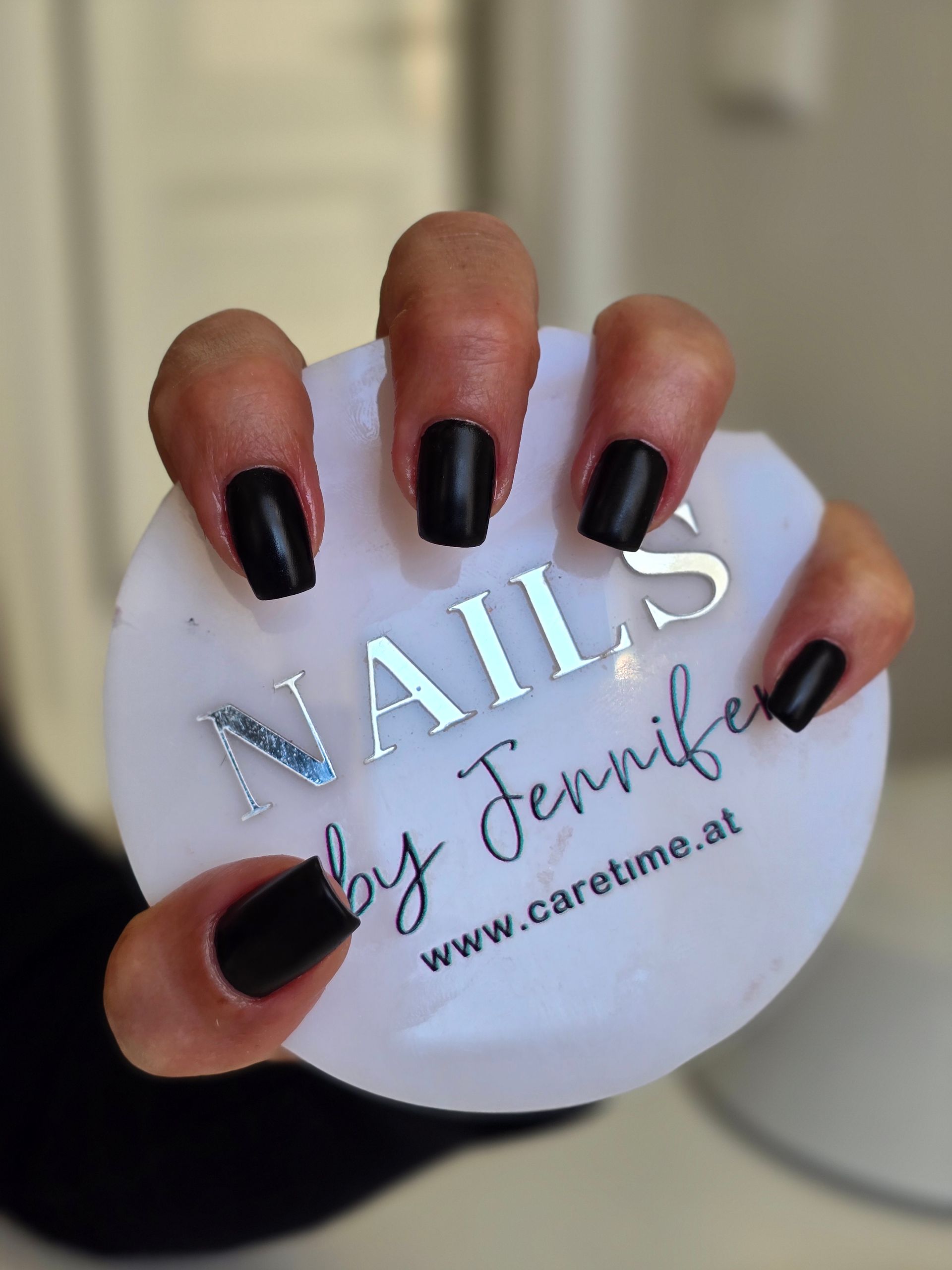 Hand mit glänzend schwarzen Nägeln, die eine runde weiße Visitenkarte eines Nagelstudios mit silberner Schrift hält.