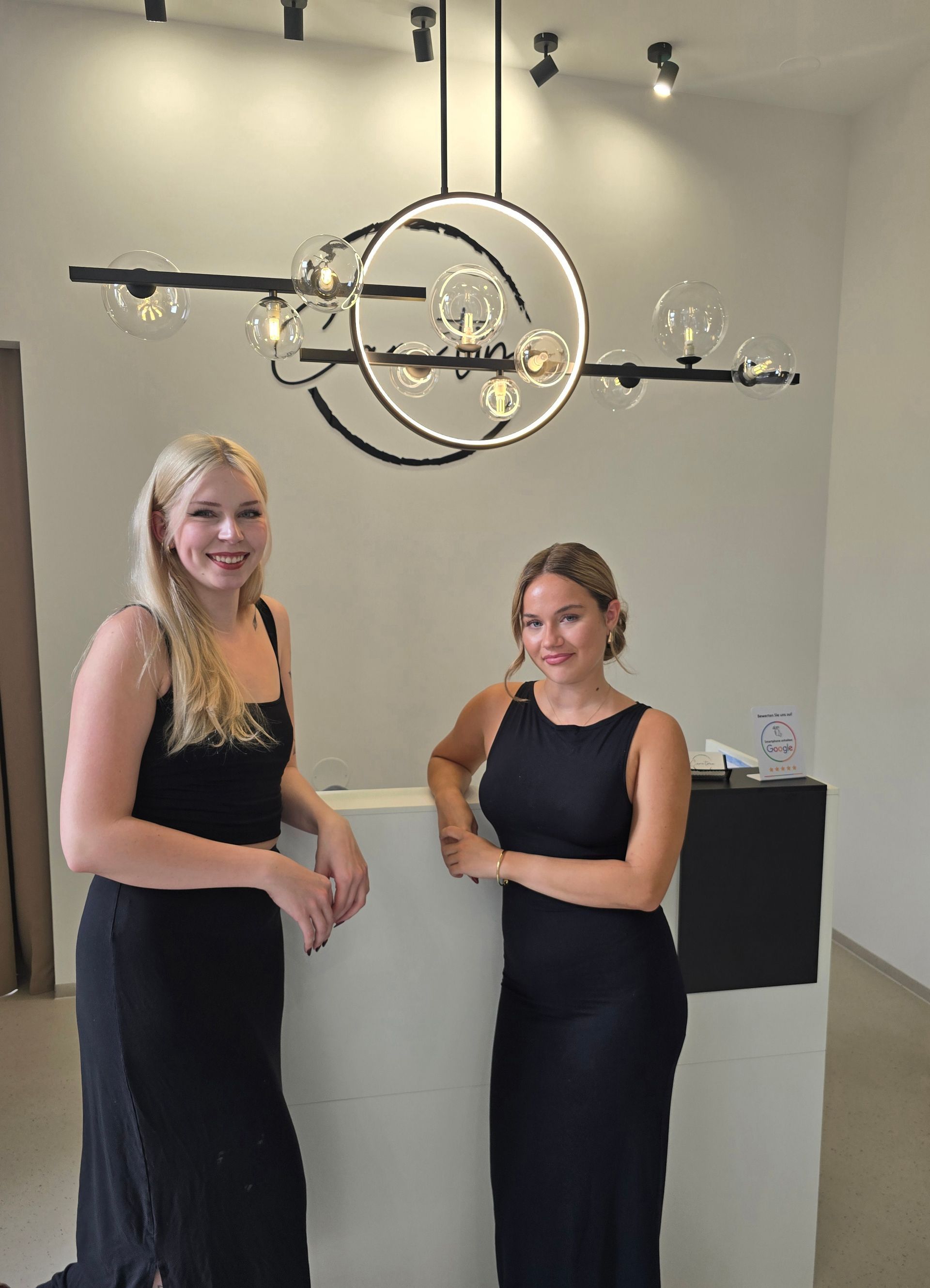 Zwei Frauen in schwarzen Outfits stehen in einer modernen Galerie unter einem runden Kronleuchter.