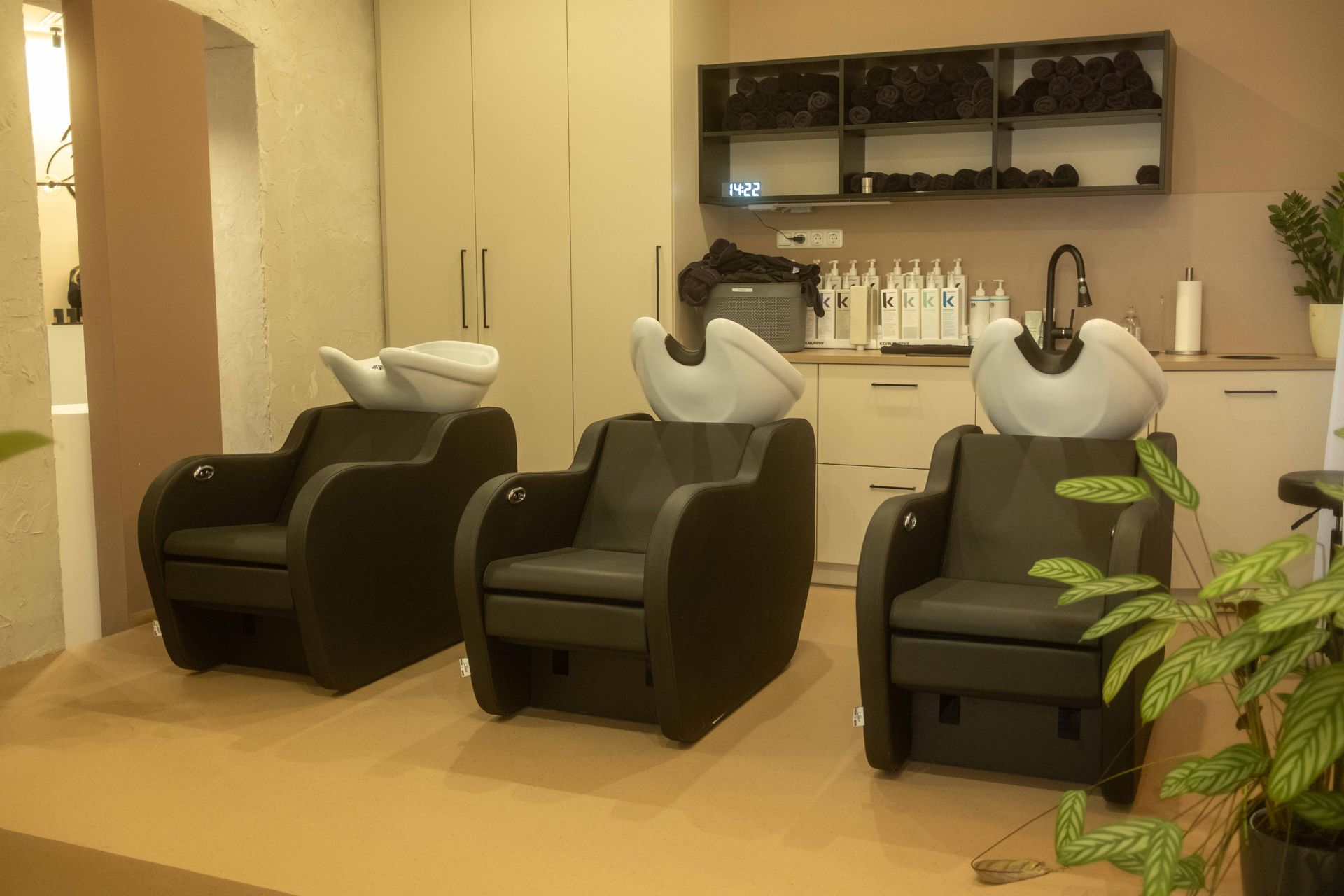 Friseurstühle und Waschbecken in einem beigefarbenen Haarwaschraum mit Schränken und Regalen.