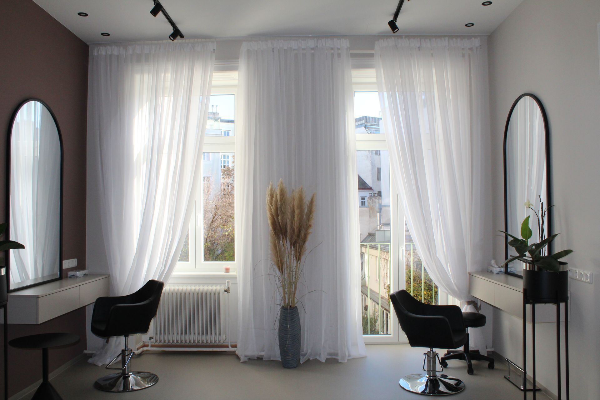 Heller, minimalistischer Salon mit zwei Friseurstühlen, Spiegeln, weißen Vorhängen und großen Fenstern.