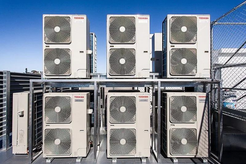 Outdoor row of rooftop air conditioning units beside a chain-link fence under a clear sky