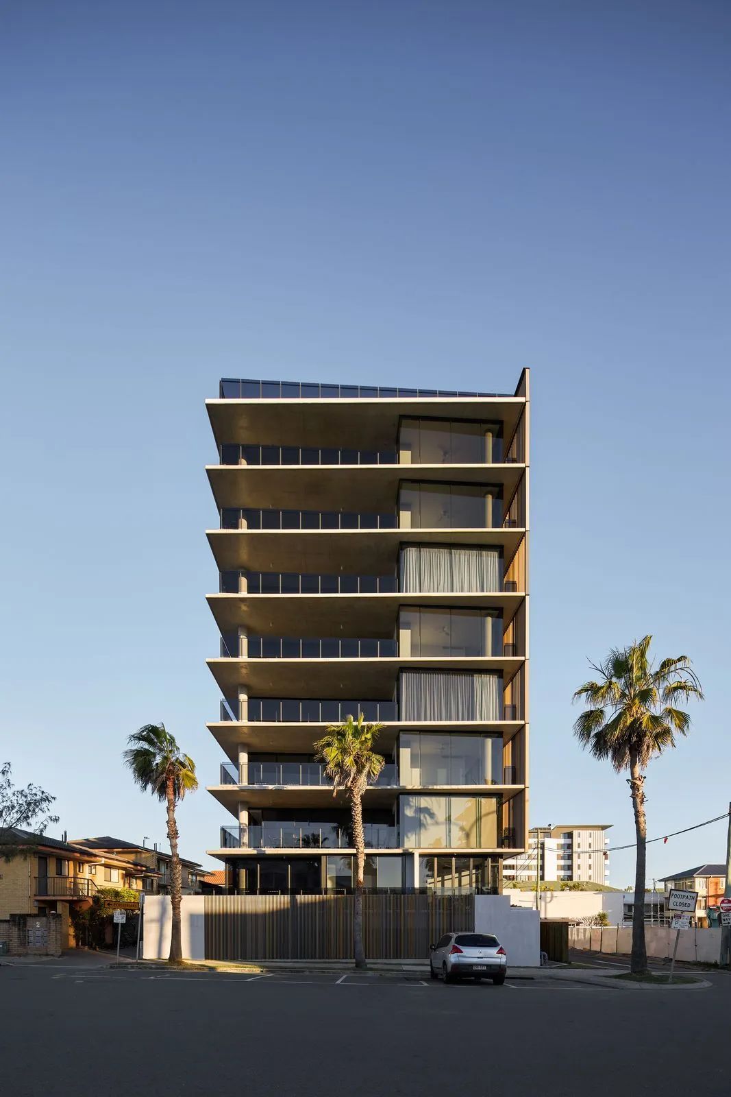 Modern mid-rise building under construction with palm trees and a parked car in front at dusk