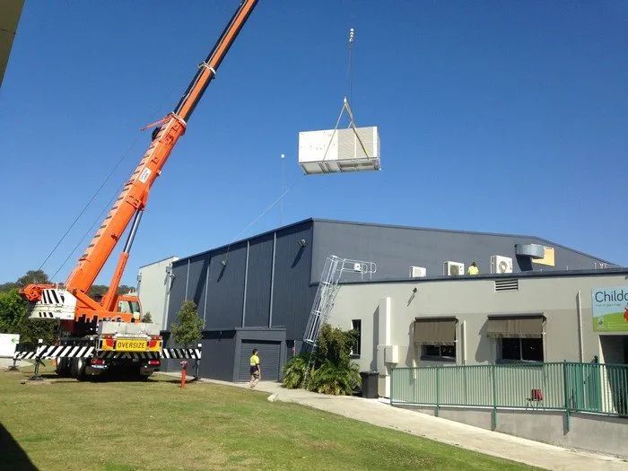 Orange crane lifting a large crate beside a blue industrial building under a clear sky