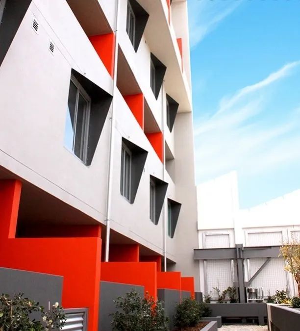 Modern building facade with red geometric accents and windows beside a courtyard under a blue sky