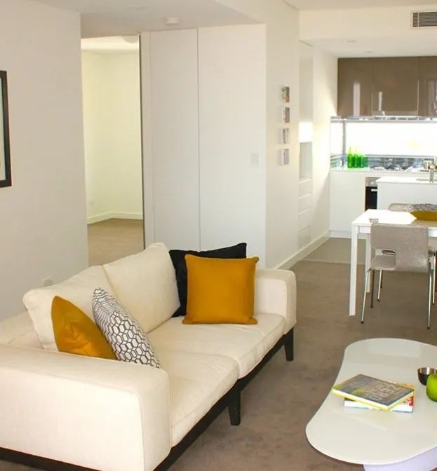 Bright modern living room with white sofa, yellow cushions, and kitchen visible in the background