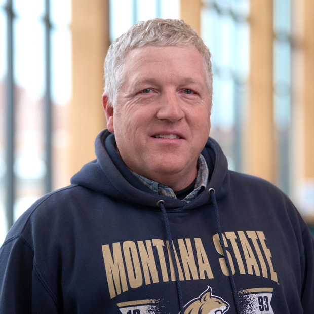 Man in a navy Montana State hoodie standing indoors, smiling softly