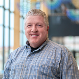 Smiling man in a blue plaid shirt standing indoors with a blurred office background