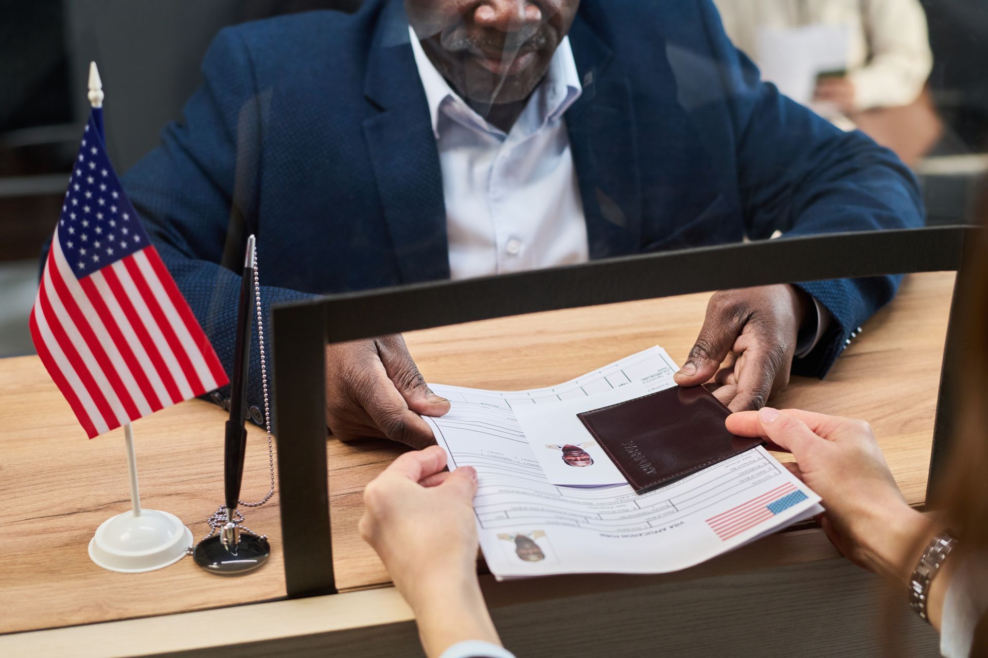 Hands reviewing a document and passport at a desk with a small U.S. flag and laptop screen.