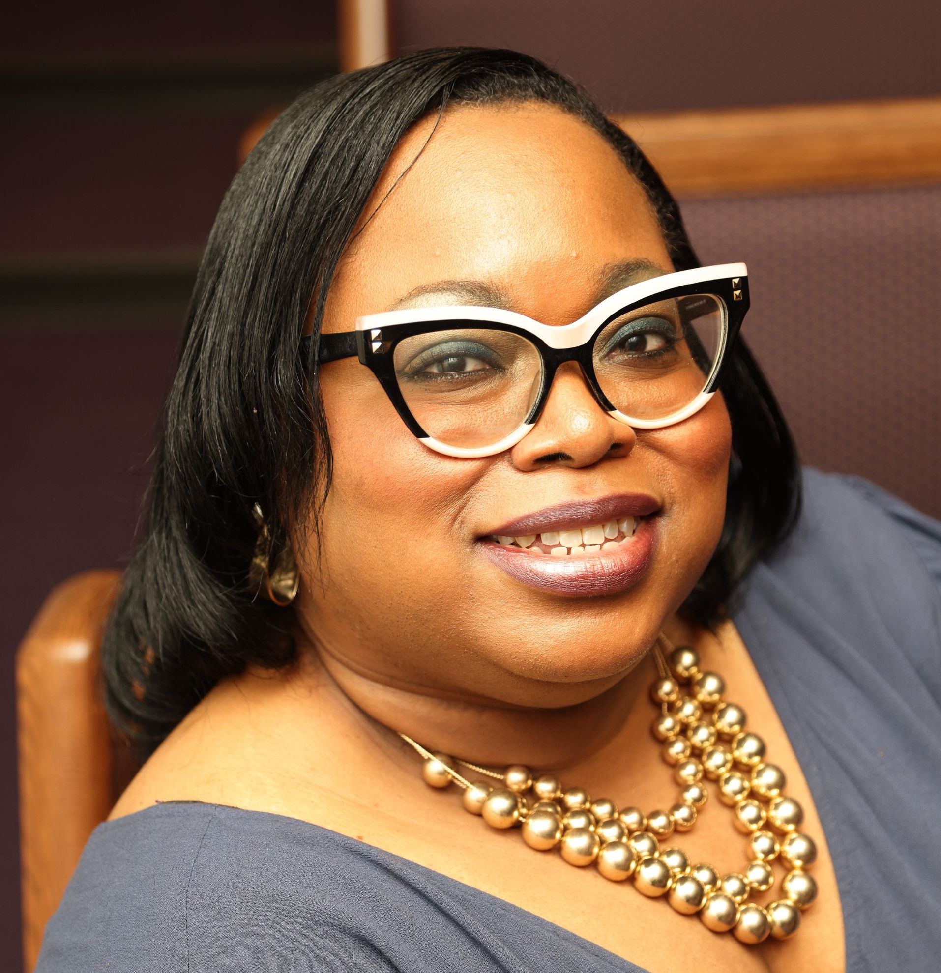 A woman wearing glasses and a silver jacket is standing in front of a green screen.