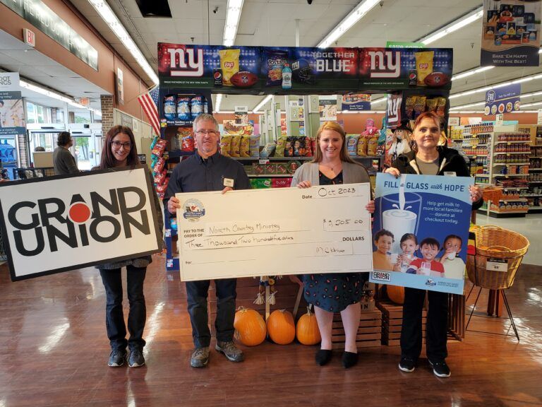 Four people pose in a store holding a large ceremonial donation check and signs for Grand Union and a milk drive.