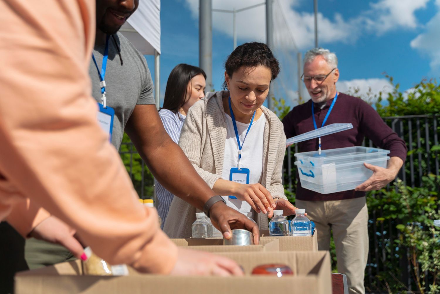 Volunteers in casual clothing organize food donations into cardboard boxes outdoors on a sunny day.