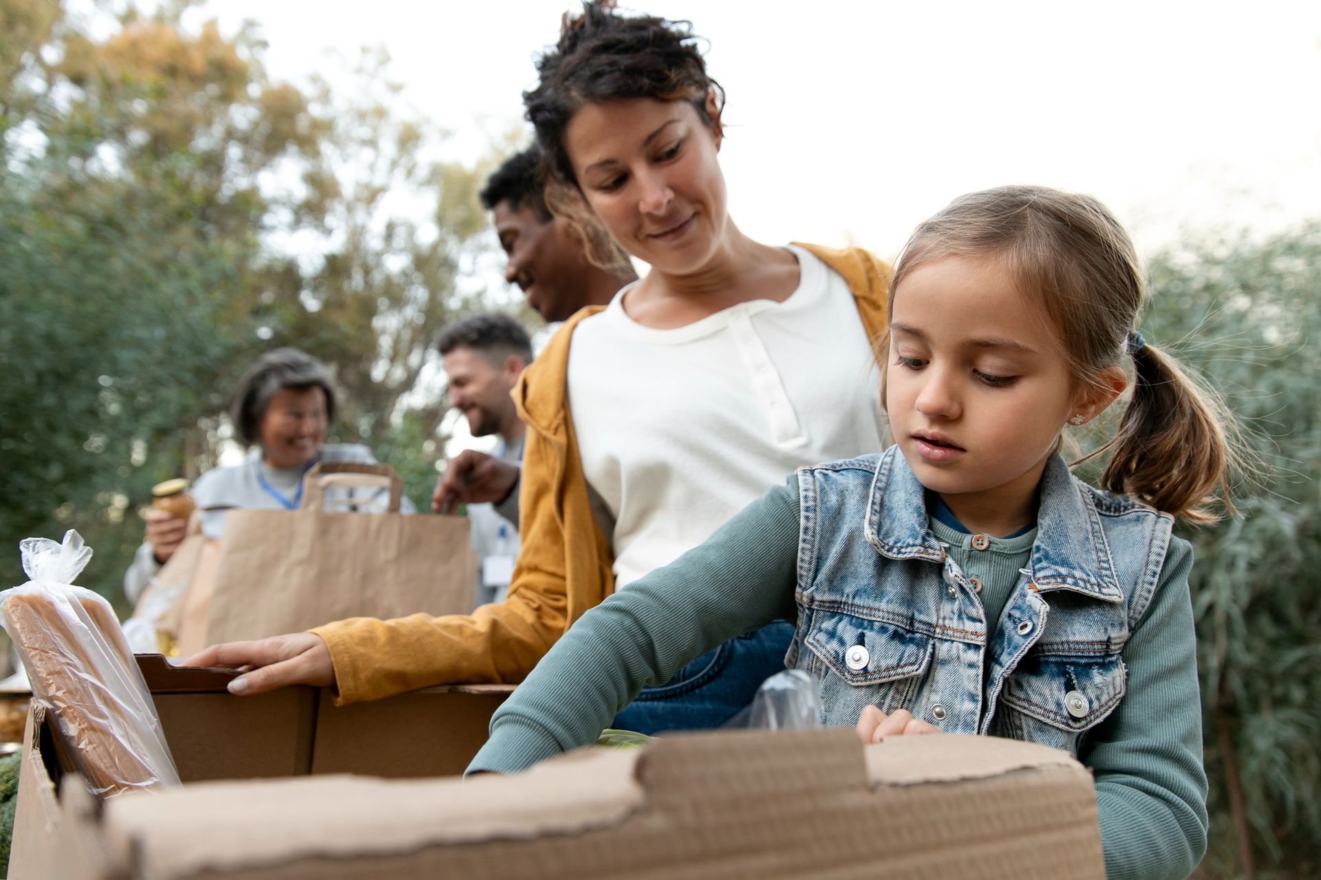 A person and a child packing food items into cardboard boxes at an outdoor volunteer event.