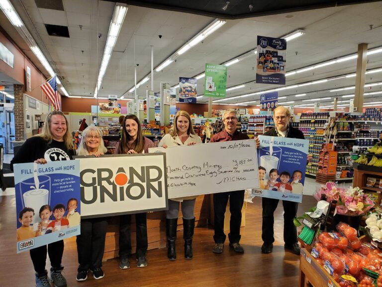 Six people hold a large Grand Union sign and a charity check inside a grocery store, representing a donation for milk.
