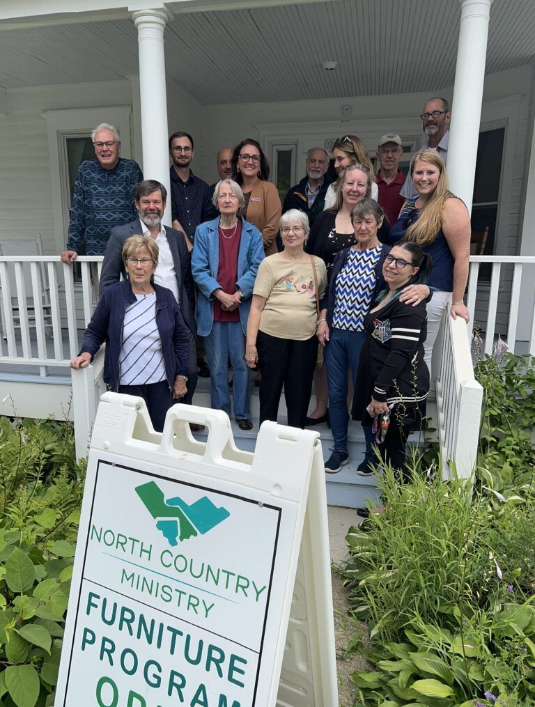 A group of people standing on a porch behind a North Country Ministry Furniture Program sign.