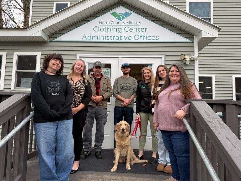 A group of seven people and a golden retriever stand smiling on the steps of the North Country Ministry Clothing Center.