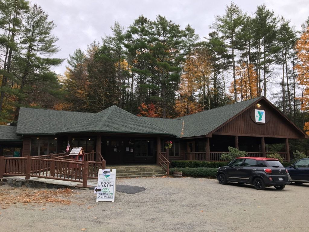 A wooden YMCA building with a green roof, set against a backdrop of autumn trees with a parked car in the gravel lot.