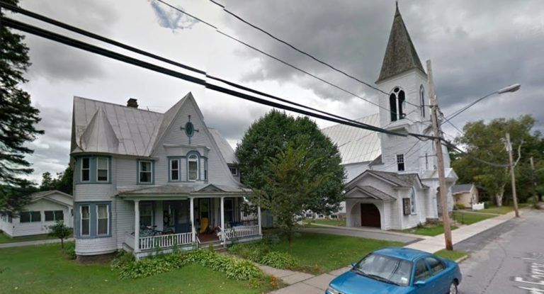 A house with a wraparound porch stands next to a white church with a tall steeple on a street with a blue car.
