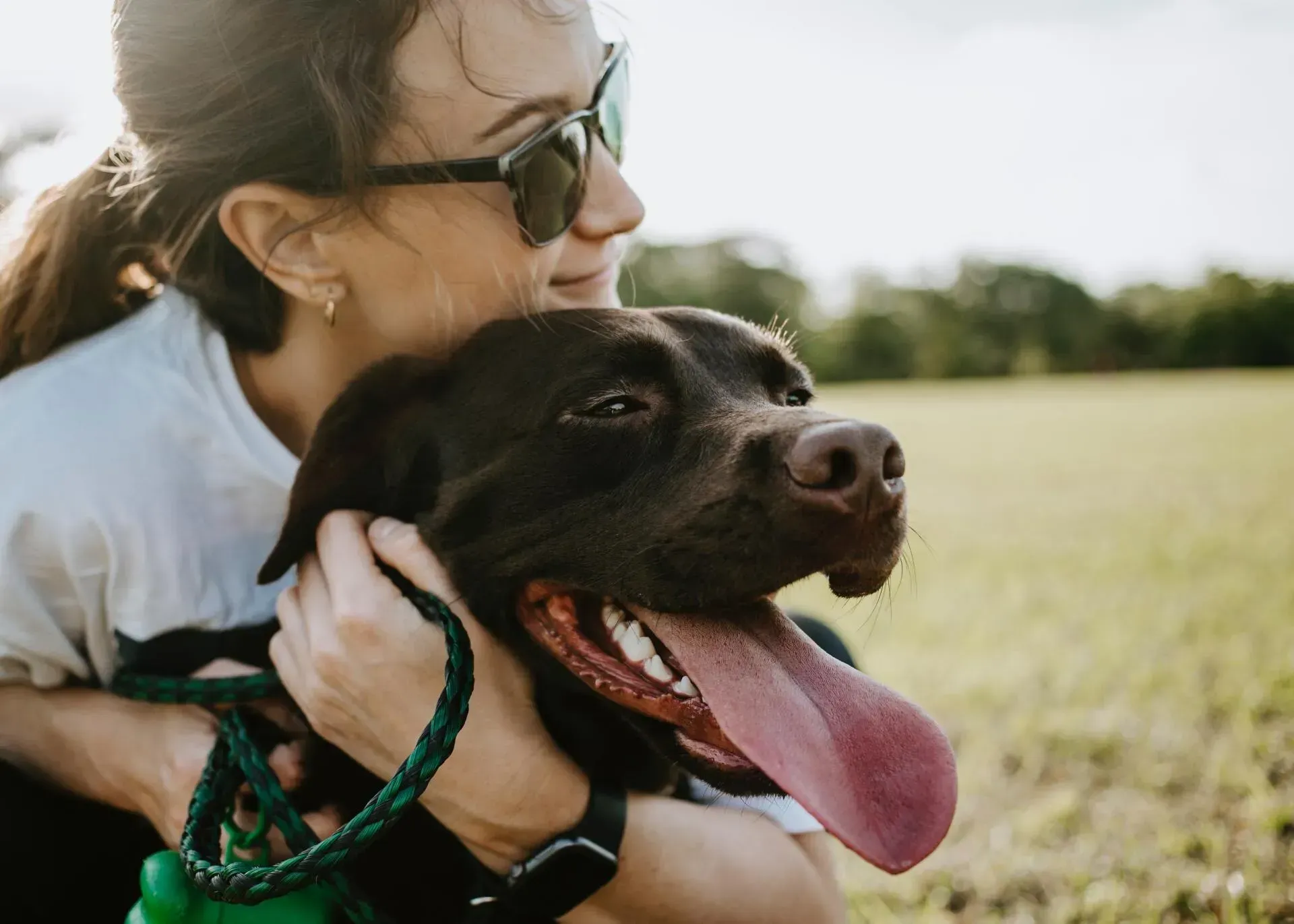 A woman wearing sunglasses hugs a happy, dark brown dog with its tongue hanging out in a grassy field.