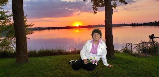 Person sitting on grass by a lake at sunset, with orange sky and trees framing the scene.