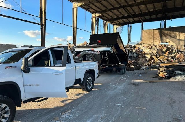 White pickup truck beside a debris pile and a burned truck under a large metal canopy in a warehouse yard