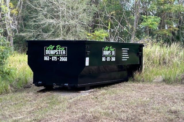 Black dumpster on grass beside trees, with white lettering and phone number on the side.