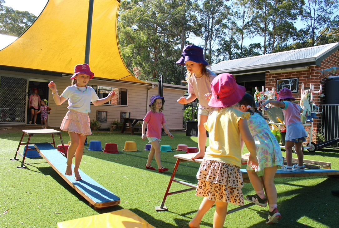 Children play on outdoor balance beams and exercise equipment in a sunny, grass-covered playground area.