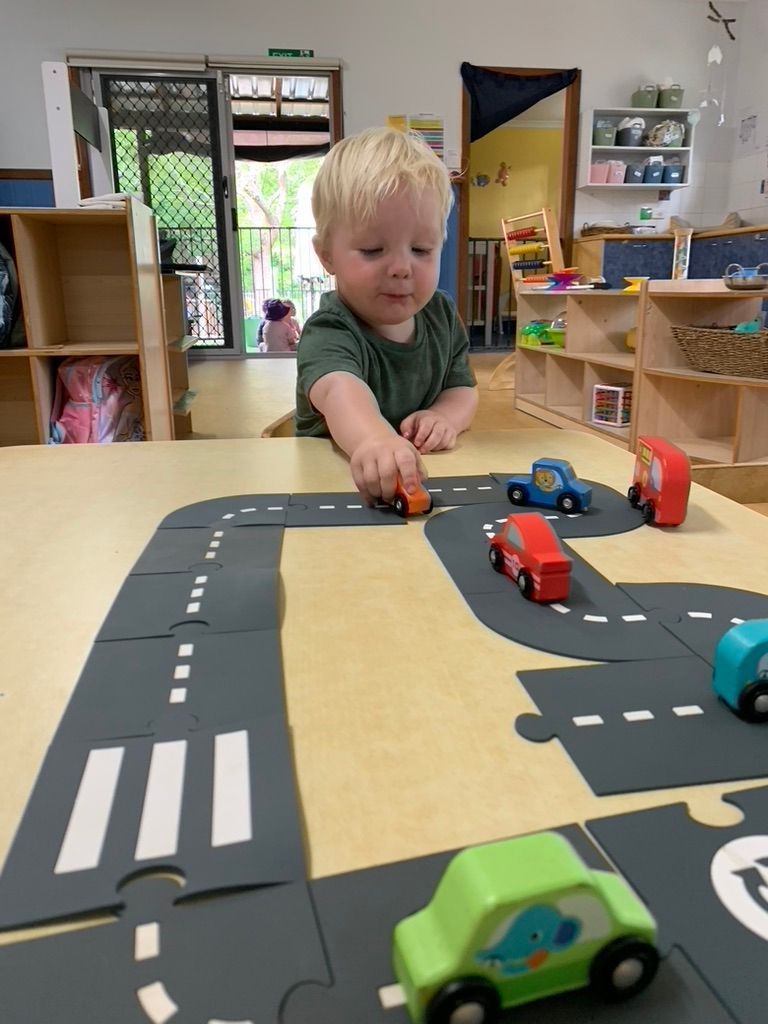 A child in a green shirt plays with small, colorful toy cars on a jigsaw puzzle road mat in a classroom setting.