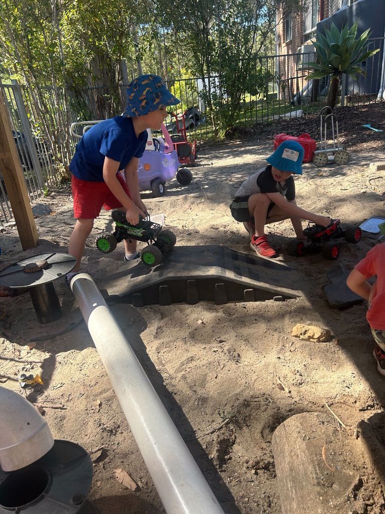 Two children wearing bucket hats play with remote-controlled toy trucks in a sandy outdoor play area.
