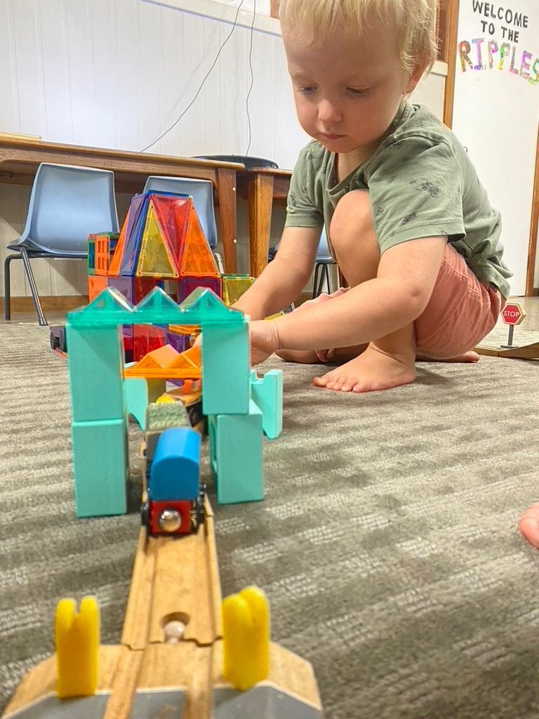 A child sits on a carpeted floor playing with a wooden toy train on tracks passing under a blue plastic block structure.