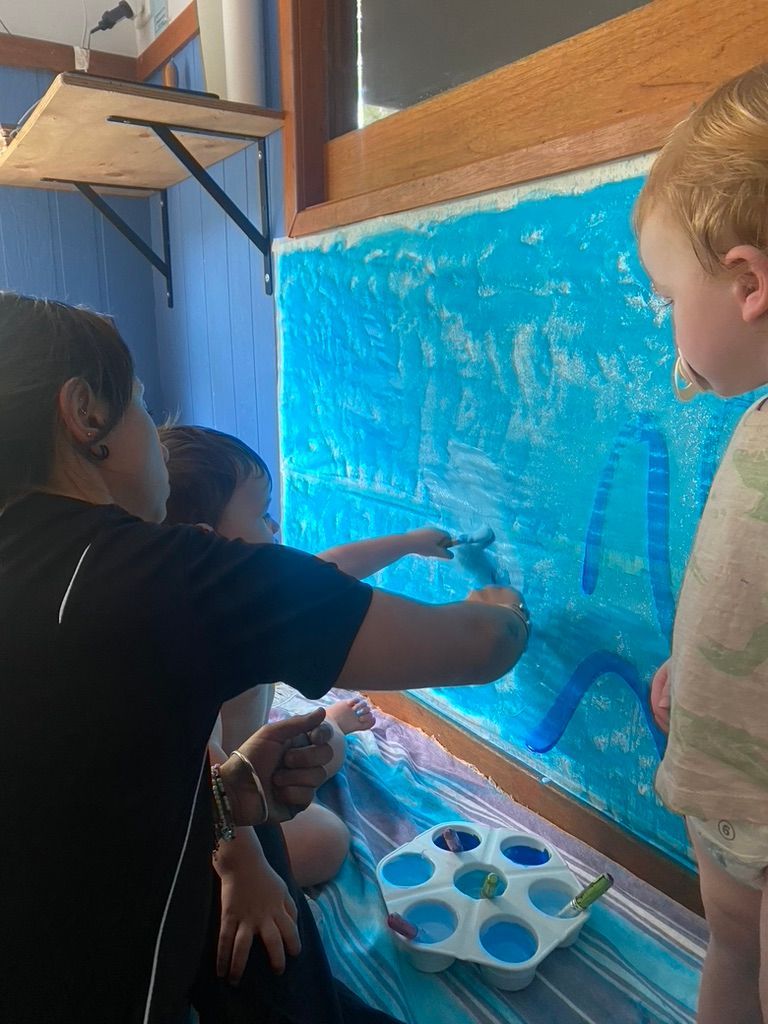 A teacher and two children paint a large blue sheet hung against a wall with brushes and a plastic palette.