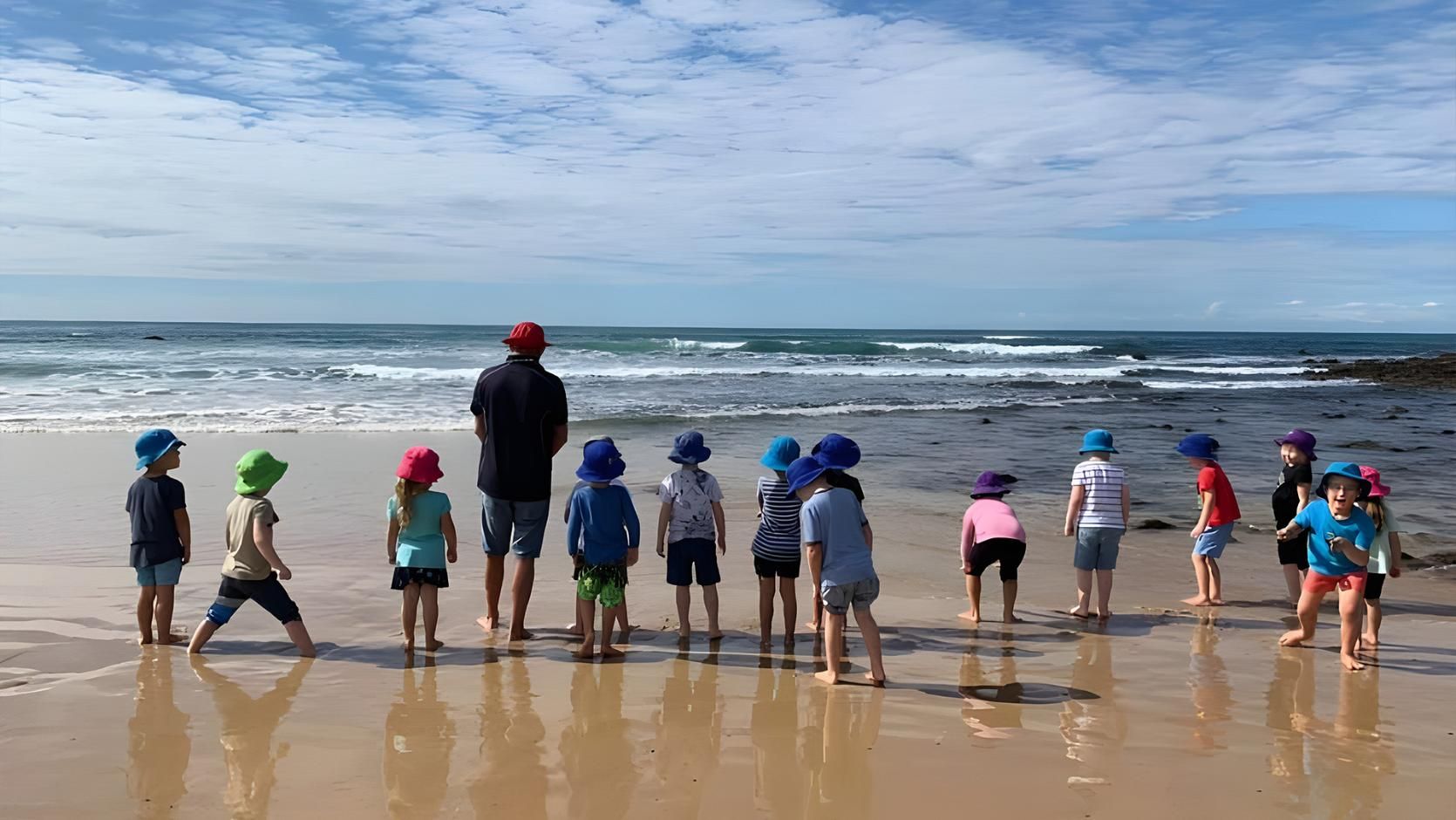 A group of children and an adult wearing hats stand on a sandy beach looking out at the ocean waves under a blue sky.