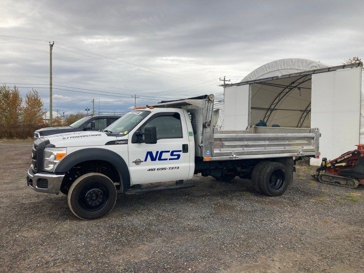 Un camion blanc avec le mot NCS sur le côté est garé sur un terrain en gravier.