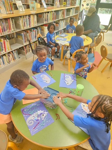 Children at a library table doing art projects. Bookshelves in the background.