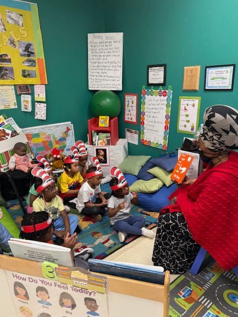 Children in Cat in the Hat hats listen to a teacher read a book in a classroom.