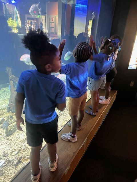 A green lizard mascot with open arms in a room with children and women watching.