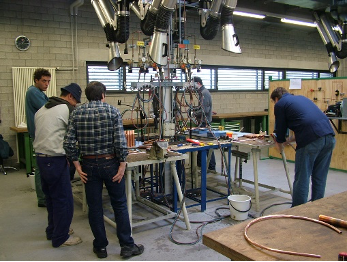 In un'officina, alcune persone si sono radunate attorno a un banco da lavoro, esaminando attrezzature e strumenti sotto i macchinari sospesi.