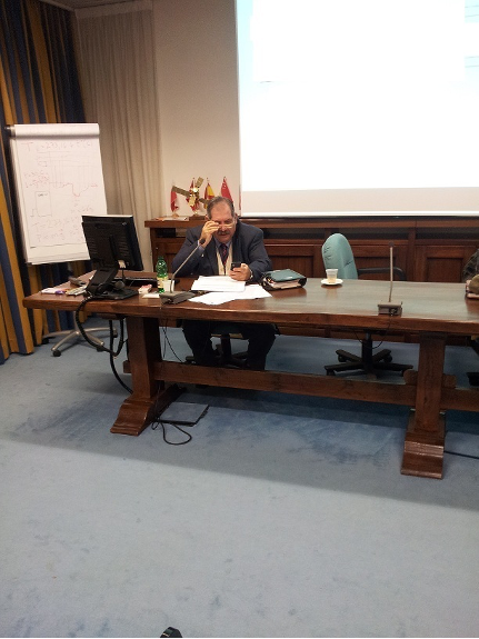 Una persona seduta a una scrivania parla in una sala conferenze, con uno schermo per proiezioni e una lavagna alle sue spalle.