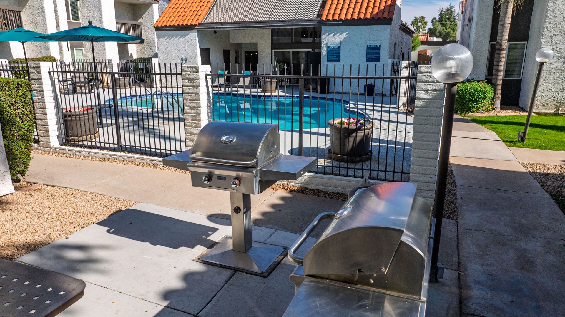 Two stainless steel grills on a patio near a pool. A fenced yard surrounds the area.