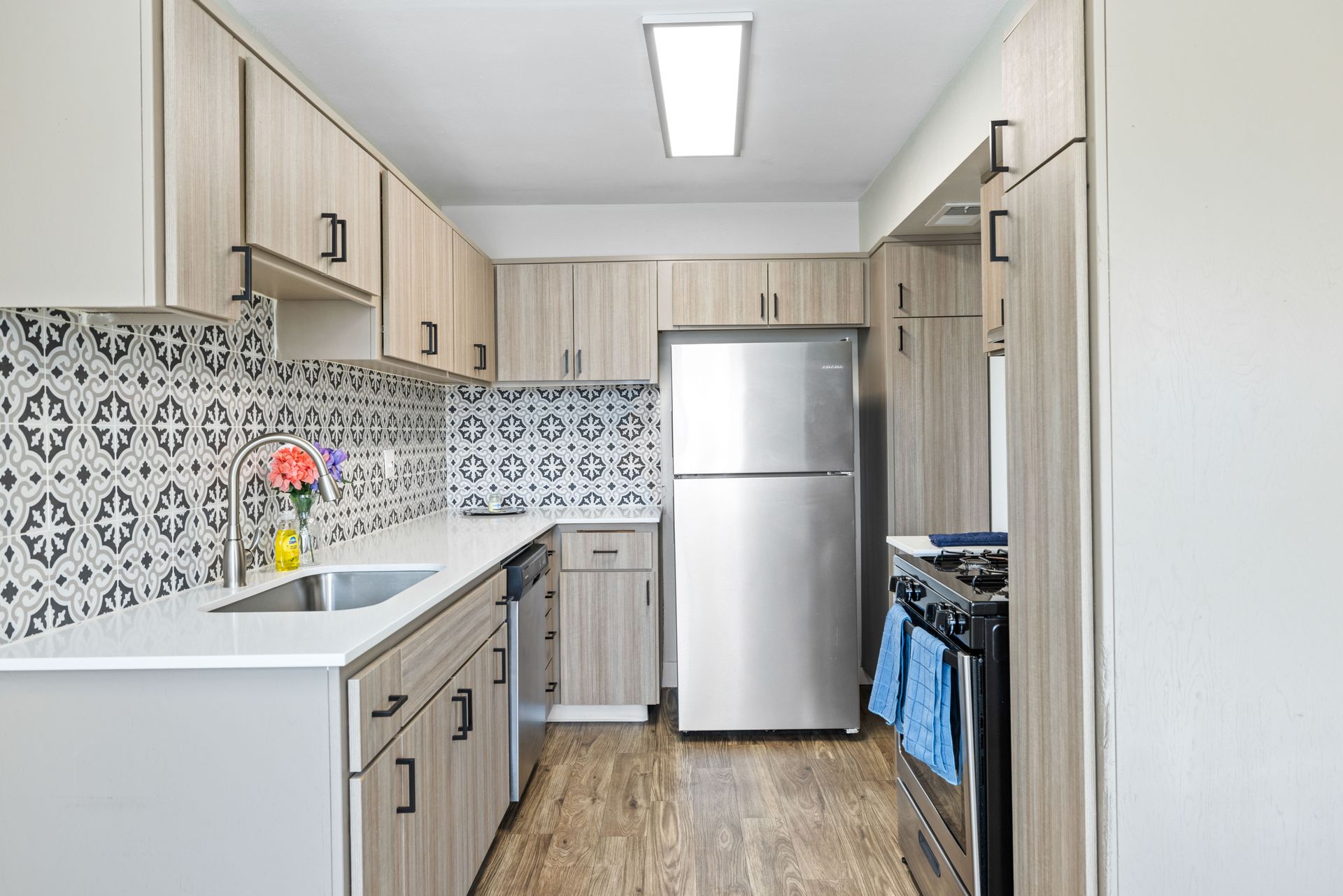 A narrow kitchen with light wood cabinets, patterned backsplash, stainless steel appliances, and wood-look flooring.