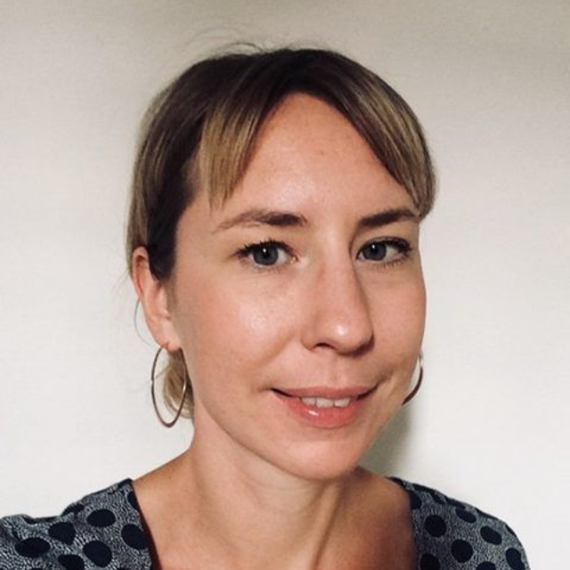 Portrait of a person with short hair, wearing hoop earrings and a patterned top, against a plain wall