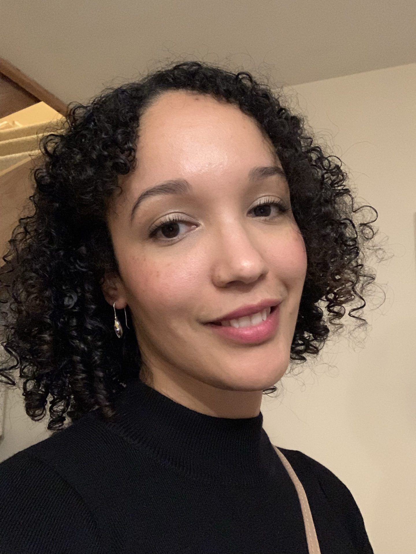 Smiling person with curly hair wearing a black top against a beige wall.