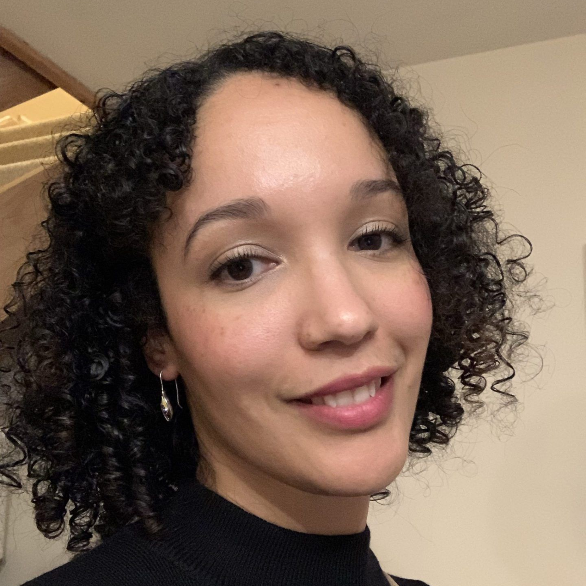 Close-up selfie of a smiling person with curly dark hair wearing a black top indoors