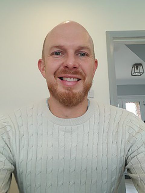 Smiling bald man in a cream cable-knit sweater indoors, standing before a light wall and doorway.