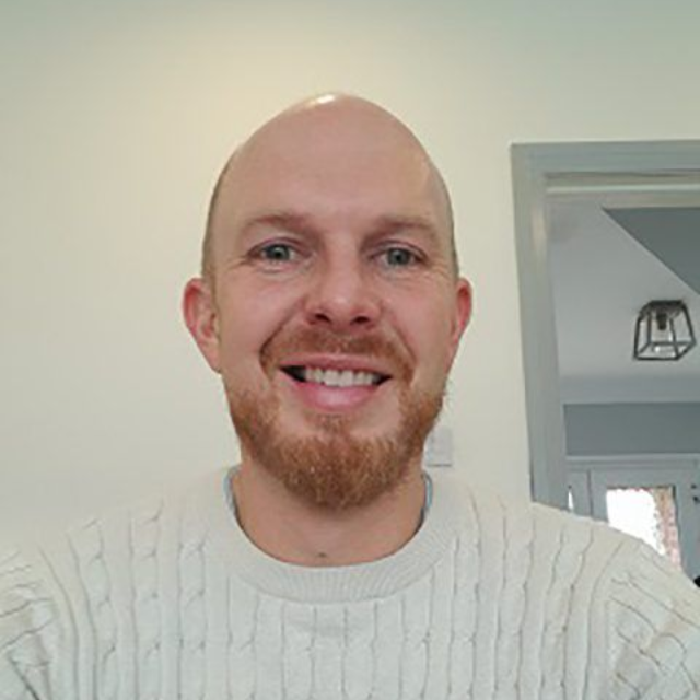Smiling bald man with a goatee in a white sweater, standing indoors near a doorway.