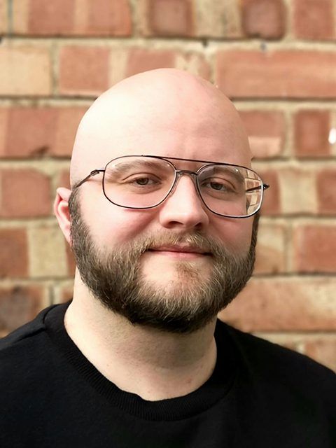 Bald man with glasses and beard smiling in front of a brick wall