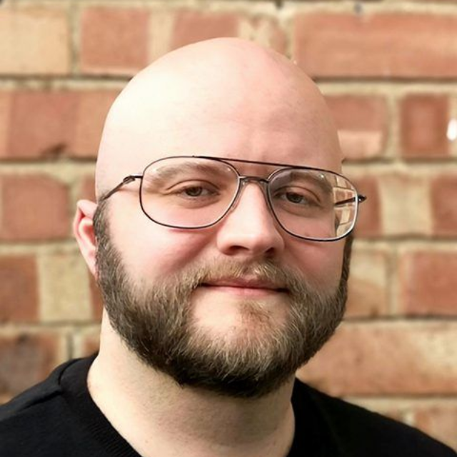 Bald man with glasses and beard, smiling in front of a brick wall