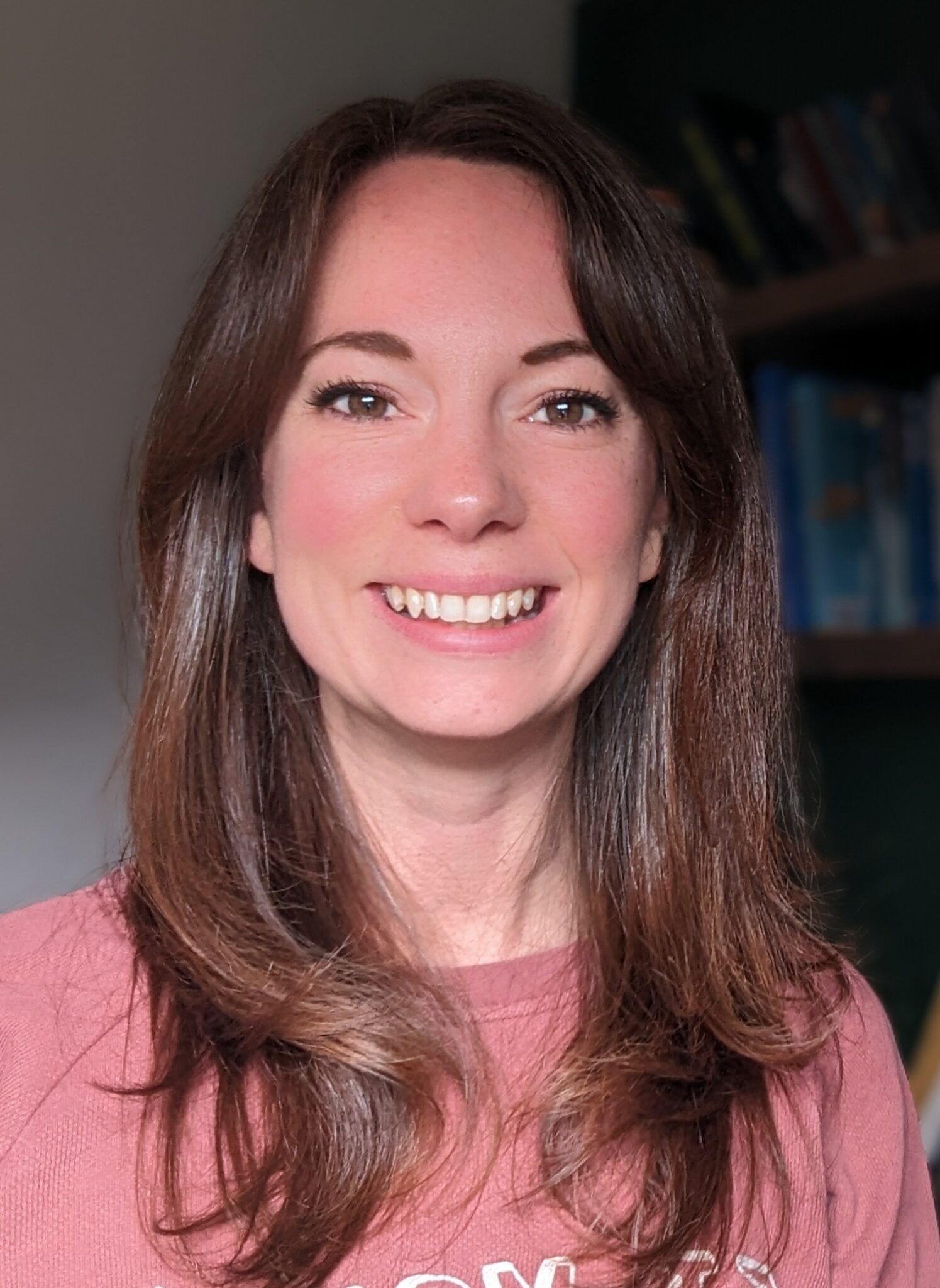 Smiling woman with long brown hair in a pink top, seated indoors against a dark background