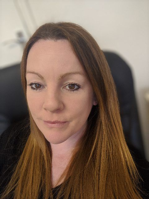 Portrait of a woman with long brown hair, wearing a black top, seated indoors against a light wall.
